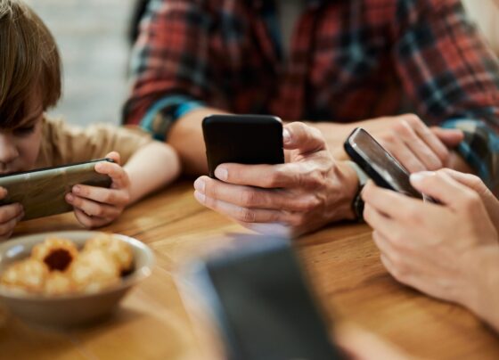 Foto colorida de pais e crianças sentados em uma mesa mexendo no celular - Metrópoles