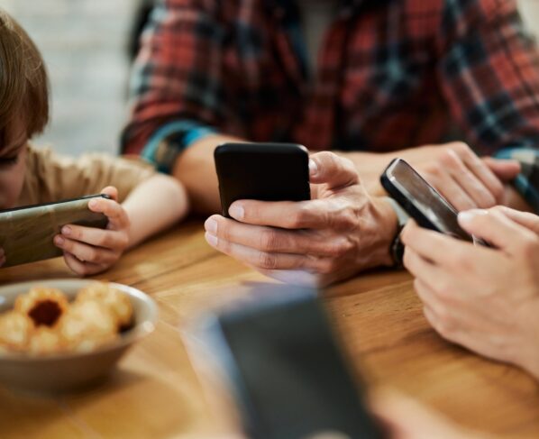 Foto colorida de pais e crianças sentados em uma mesa mexendo no celular - Metrópoles
