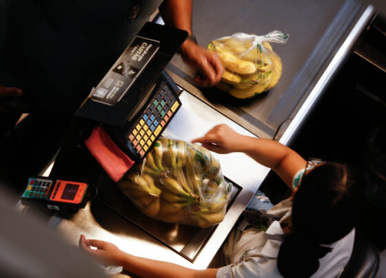 Imagem colorida mostra mulher pesando banana no caixa de mercado - Metrópoles