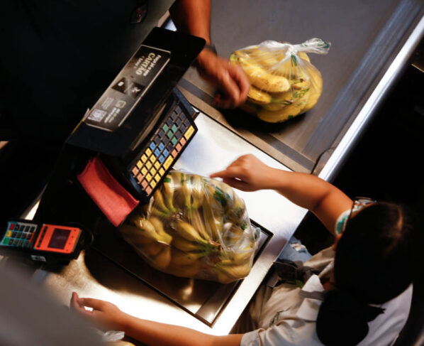 Imagem colorida mostra mulher pesando banana no caixa de mercado - Metrópoles